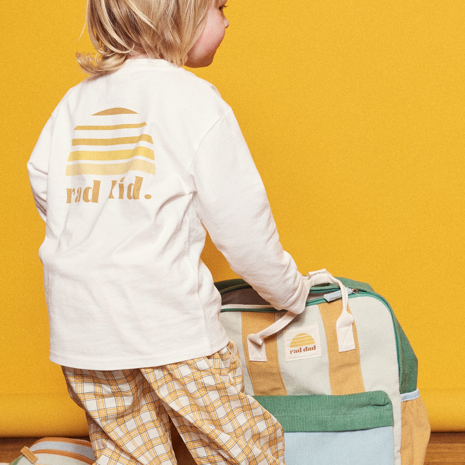 Child wearing a white long sleeve top with 'rad kid' text, standing next to a yellow,green and blue backpack on a yellow background