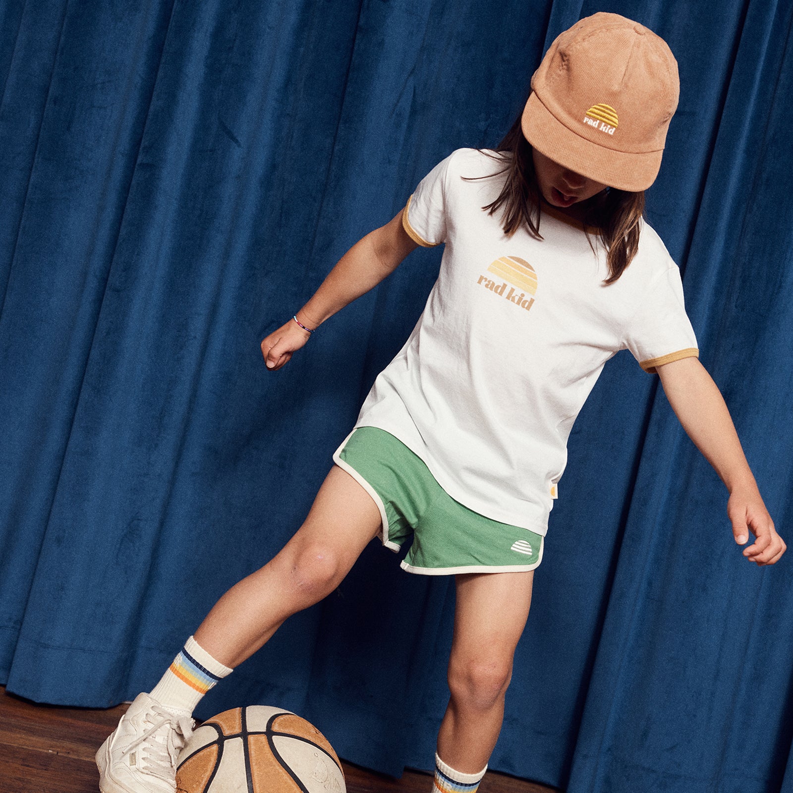 Person wearing a white t-shirt and green shorts with a brown cap, standing next to a basketball against a blue curtain.