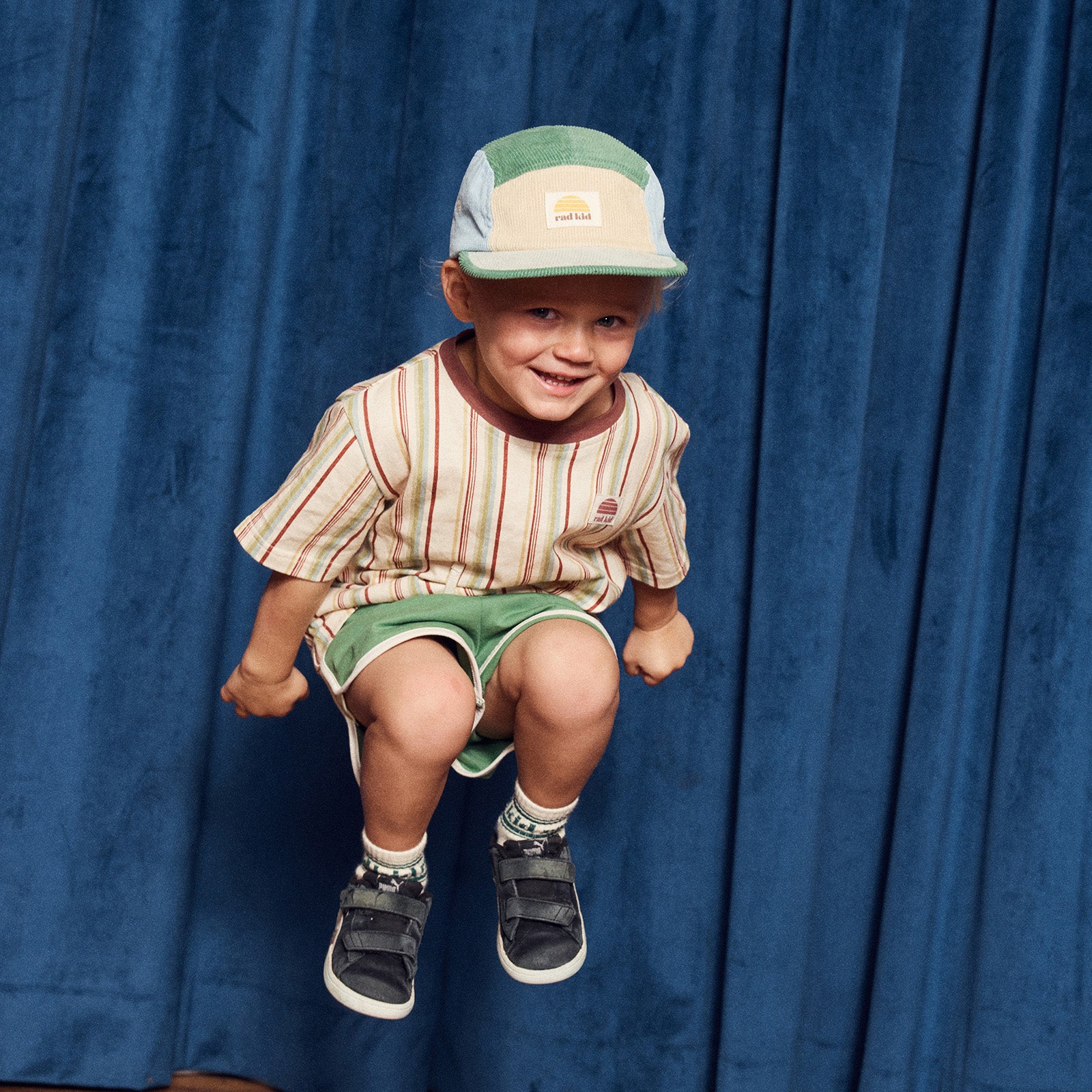 Child wearing a striped shirt, green shorts, and a cap against a blue curtain background