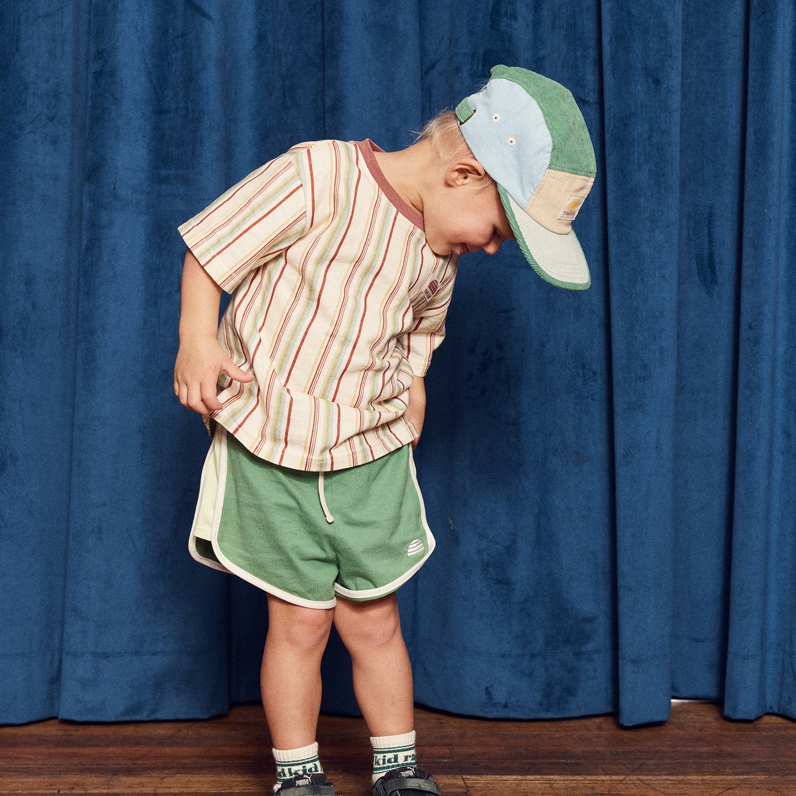 Child wearing a striped shirt and green shorts with a cap, standing against a blue curtain.