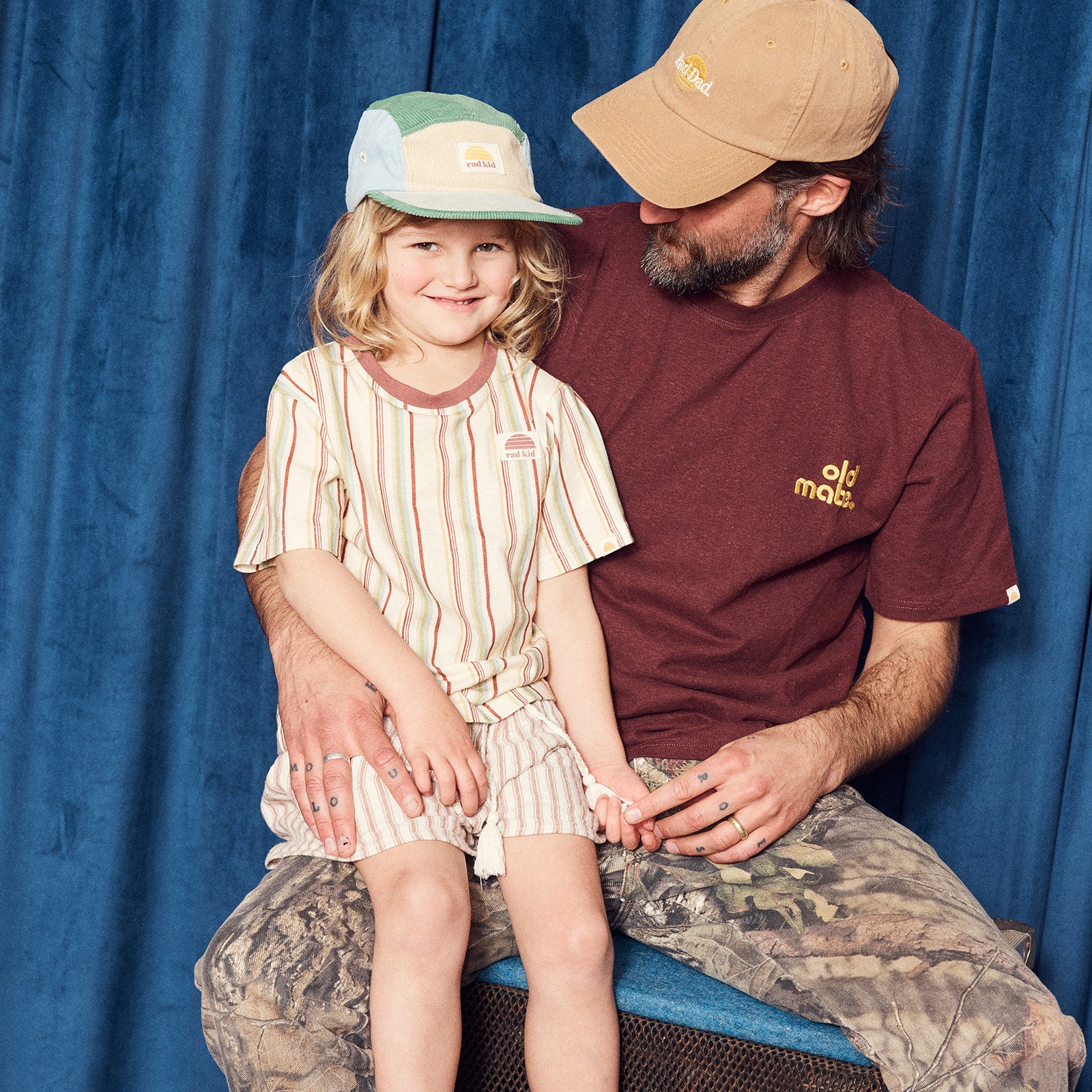 Man and child sitting together wearing hats and shirts against a blue curtain background.