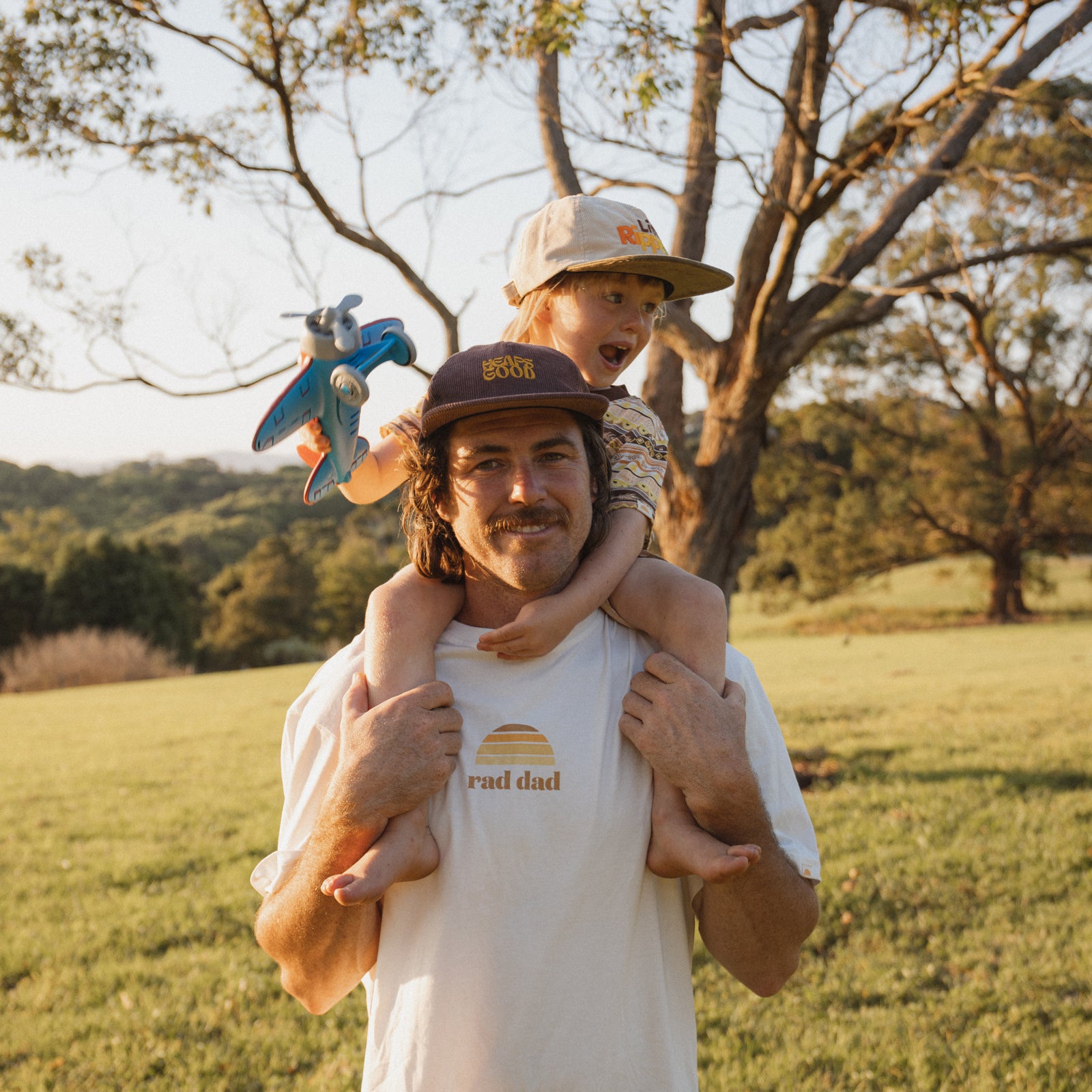Man wearing white "Rad Dad" tee and "Heaps Good" cap with child on his shoulders wearing "Little Ripper" cap