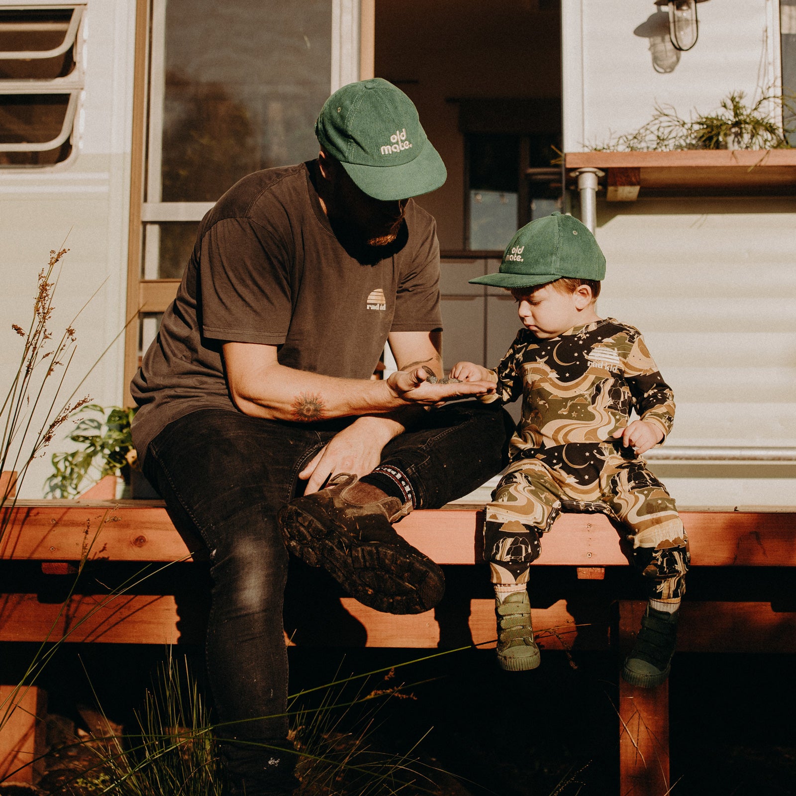 Man and child sitting on a bench in front of a trailer, both wearing green caps.