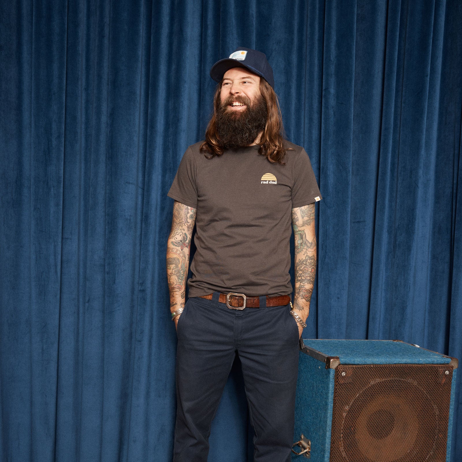 Man with tattoos and a beard wearing a trucker cap and "rad dad" t-shirt, standing next to a speaker against a blue curtain.