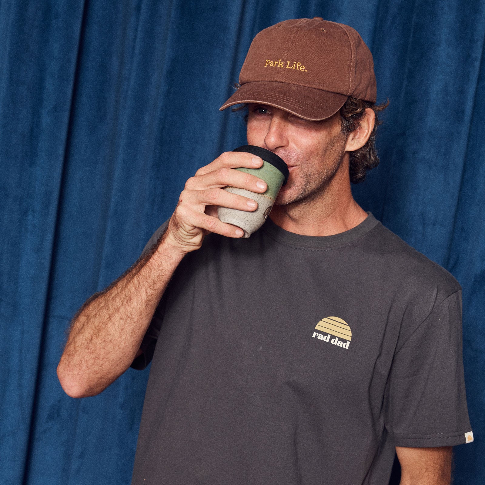Man wearing a brown cap and dark t-shirt with logos, drinking from a green cup against a blue curtain background.