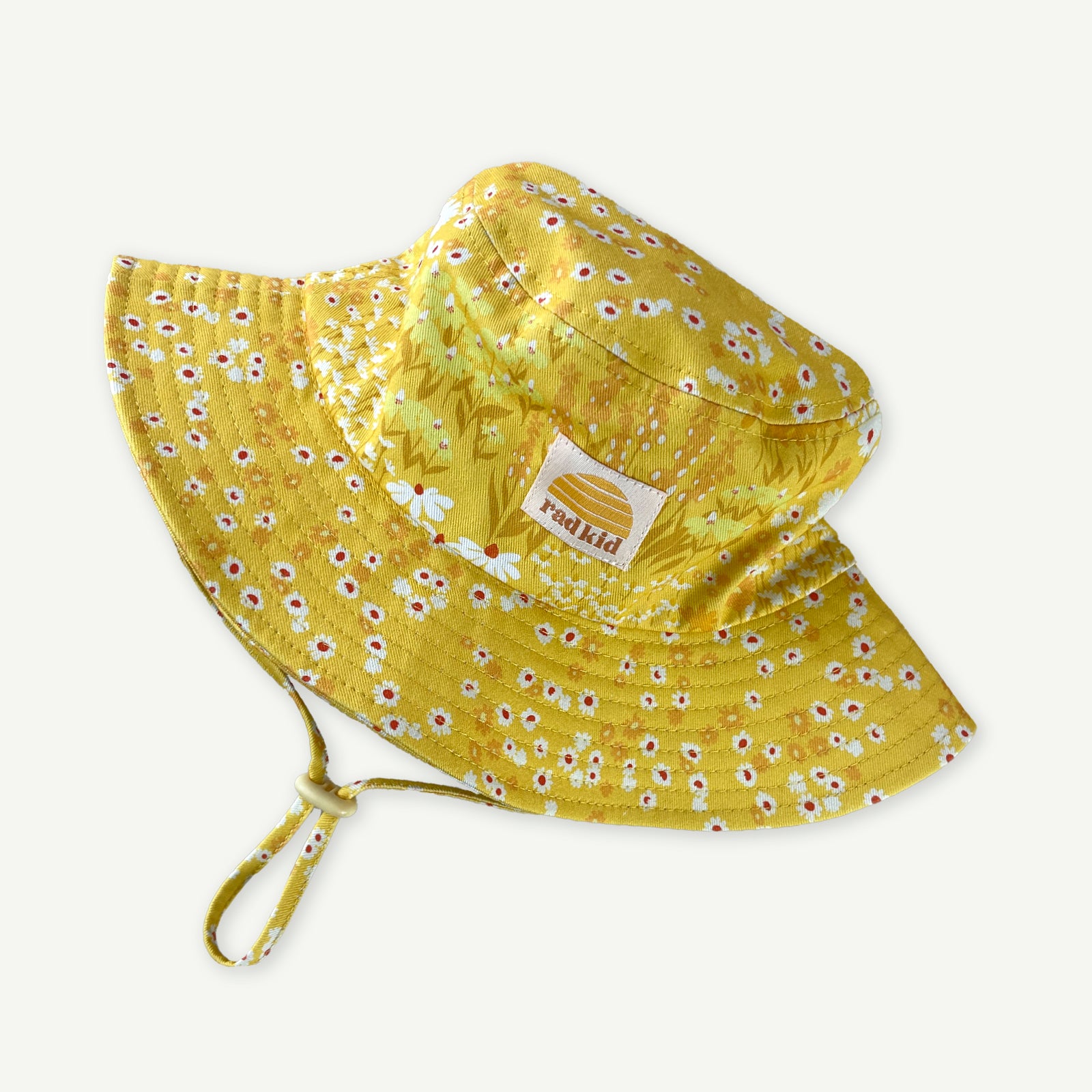 Bucket hat in a yellow Buttercup meadow of flowers print with Rad Kid patch in off white with Banabae sun Logo in Yellow sewn to Centre front, drawstring in print with yellow strap adjusted.