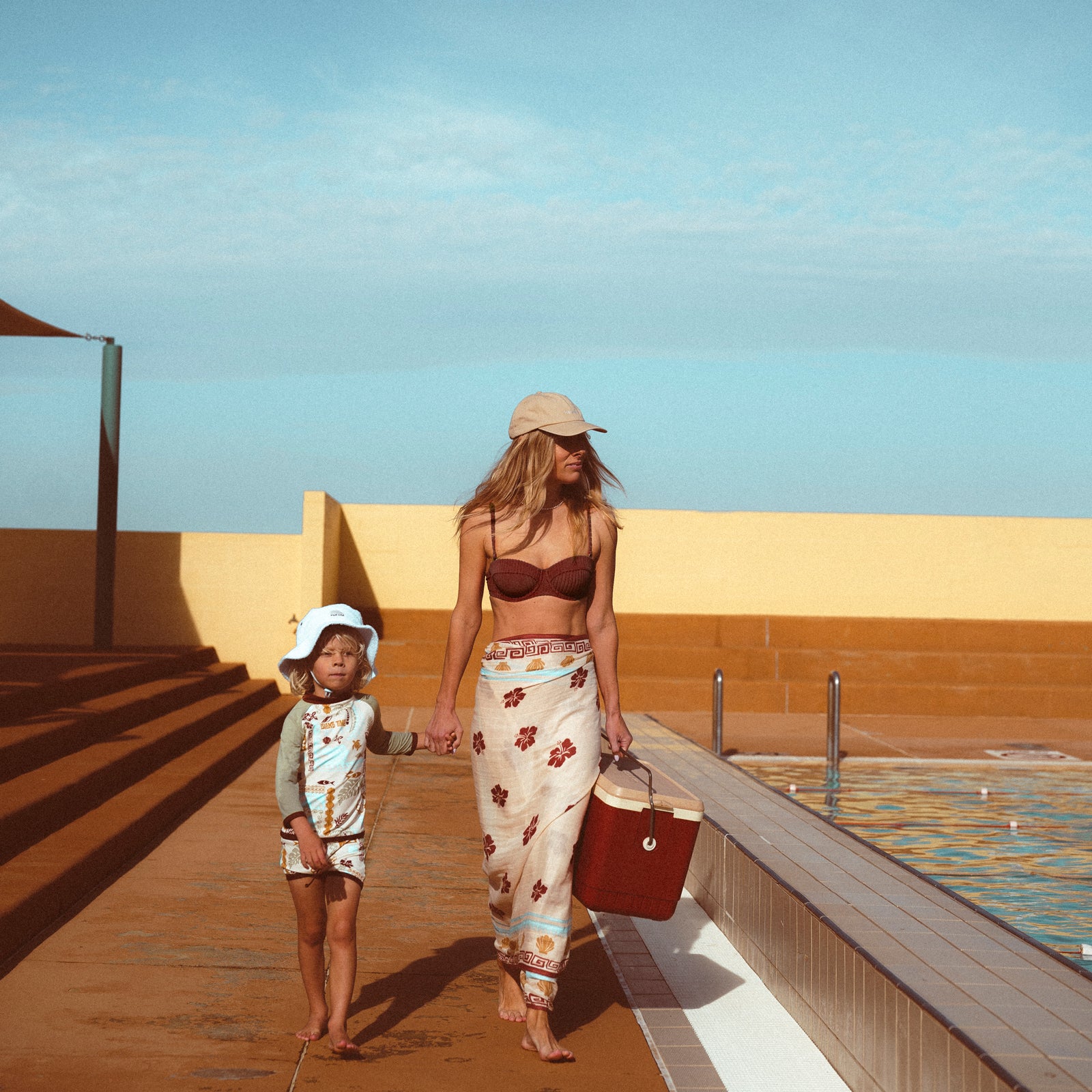 Lifestyle image of mother and son at an open air pool walking together holding hands beside the pool. The mother is wearing the Banabae washed sand cap in organic cotton twill, Banabae embroidery at the center front in contrast white. Brown bikini and Banabae Island Time Sarong. Child is wearing Rad Kid Toweling bucket hat in soft blue and Island Time rash vest and shorts.
