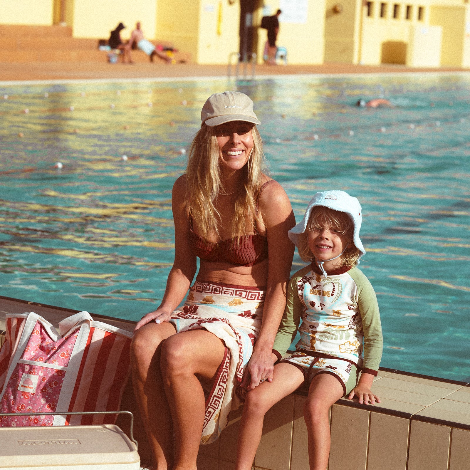 Lifestyle image of mother and son at an open air pool sitting together at the side of the pool. The mother is wearing the Banabae washed sand cap in organic cotton twill, Banabae embroidery at the center front in contrast white. Brown bikini and Banabae Island Time Sarong. Child is wearing Rad Kid Toweling bucket hat in soft blue and Island Time rash vest and shorts.