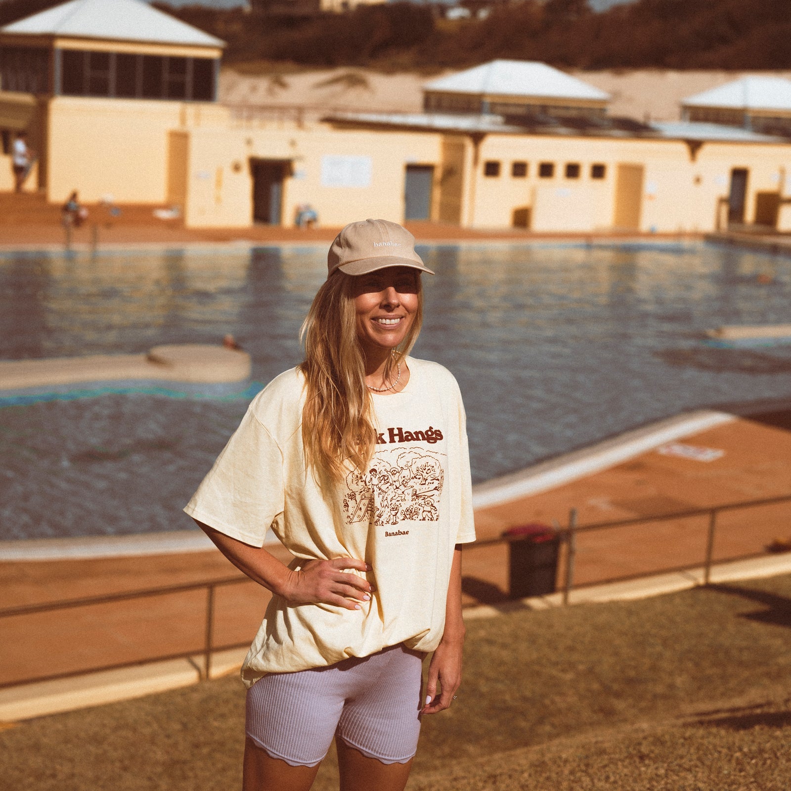Lifestyle image of Woman wearing the Banabae washed sand cap in organic cotton twill, Banabae embroidery at the center front in contrast white. Oversized Park Hangs Tee in yellow with brown print and lilac ribbed bike shorts. Standing in front of a pool.