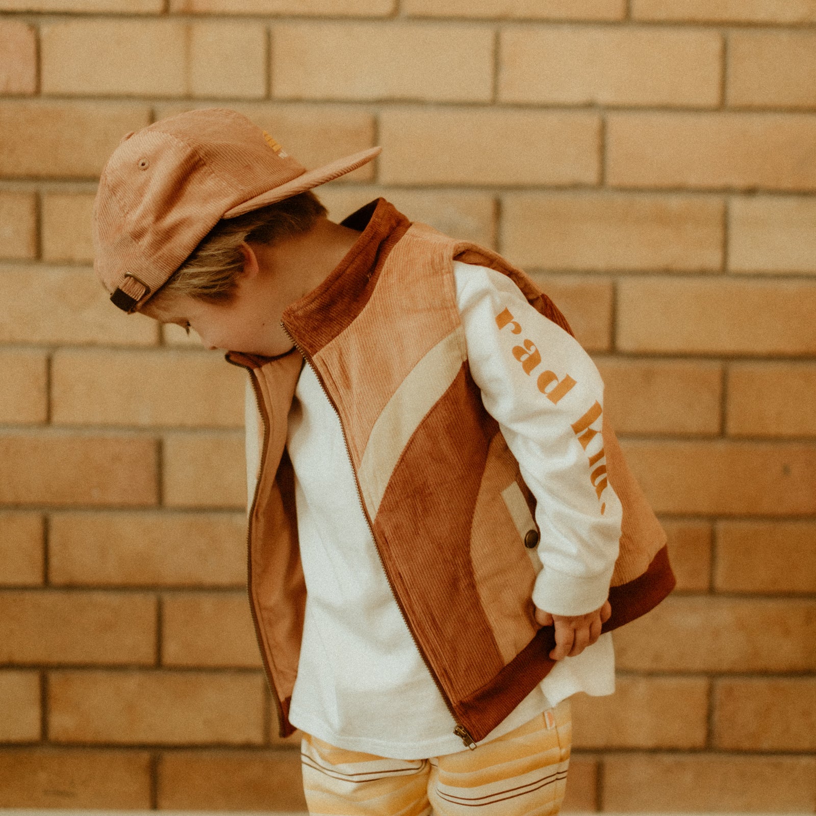 child in long sleeve 'rad kid' tee with tan cord vest over it and backwards tan cap