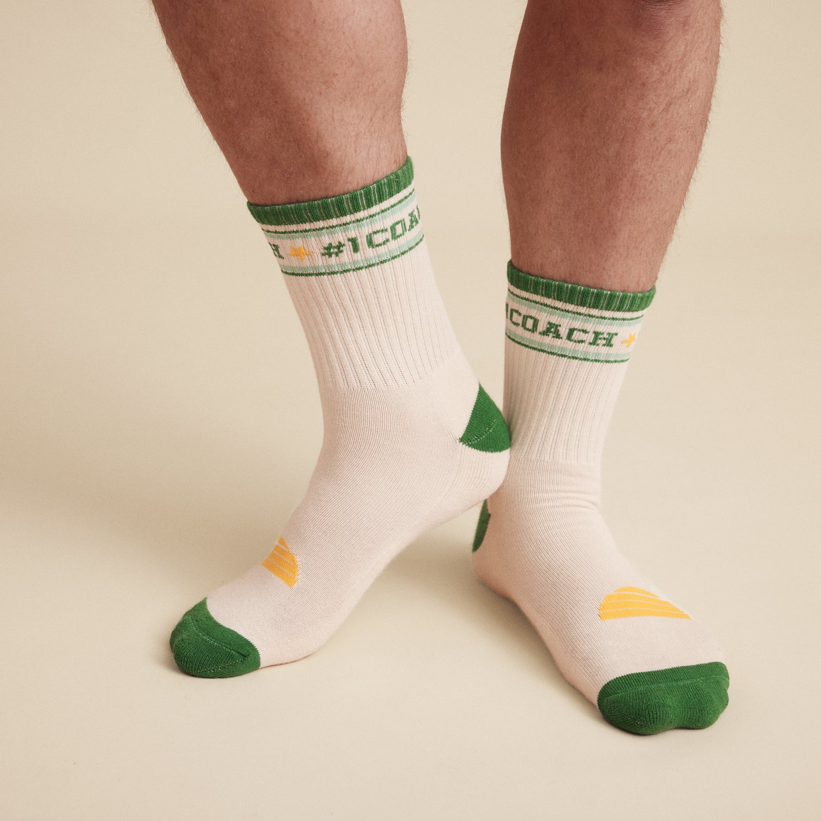 close up Image of socks worn on the body. #1 Coach socks, White crew socks with #1 coach in Green written horizontally, bordered with a green and aqua stripe. Toe and heal pads in contrast green with Banabae written along the sole in yellow, and the iconic Banabae sun logo in yellow placed on top of the foot when worn.