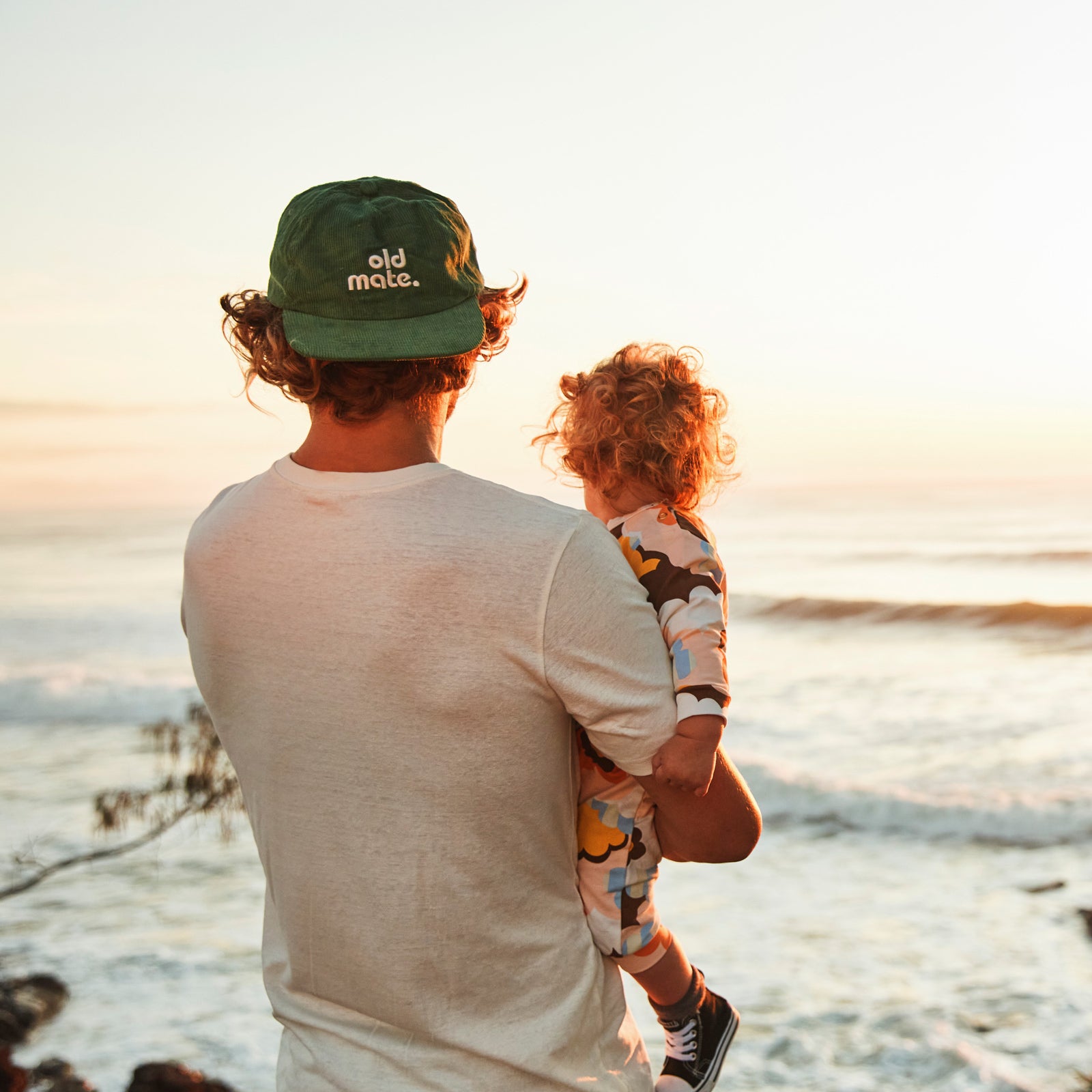 Lifestyle image of the back view of a man holding a small child looking out at ocean. the man has the Old Mate Cap on backwards . Cap is Moss green corduroy pinch front cap with old mate embroidered on center front in contrast off white and a white T-Shirt.