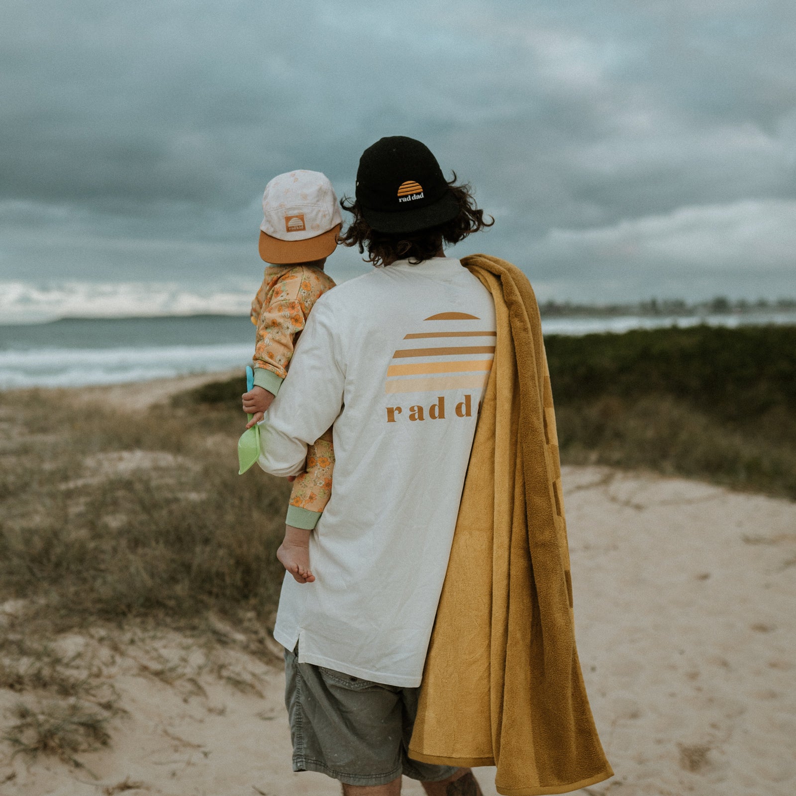 man holding child both wearing caps and the dad wearing a 'rad dad' long sleeve white tee