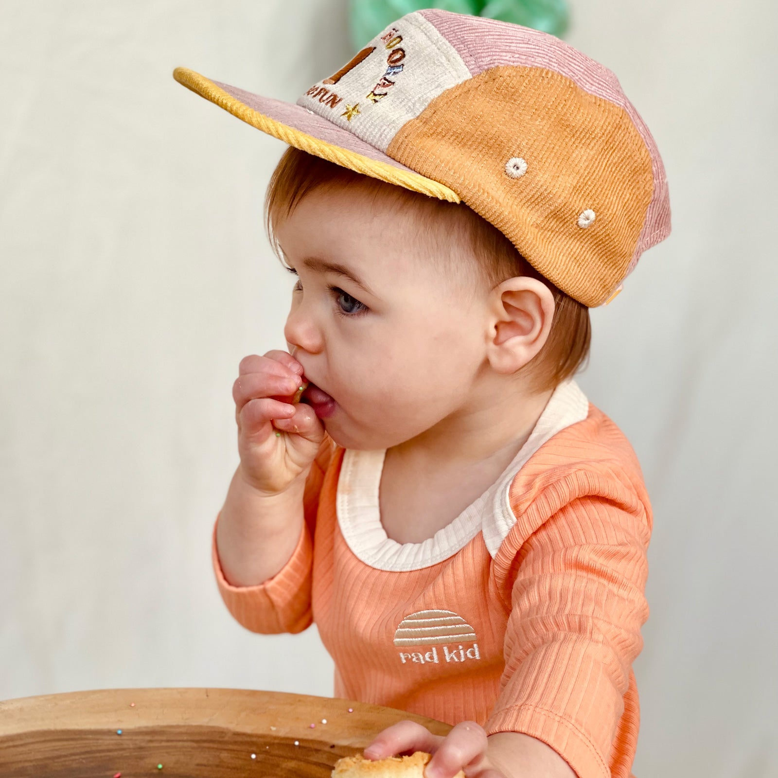 Party time photo, one year old child wearing Banabae Peach Ribbed Organic Cotton Long Sleeve Onesie with contrast natural neck bind and Rad kid/Banabae Sun Logo embroidered on right hand . Child is also wearing the 1st Birthday Cord Cap musk Spliced, she is looking super cute eating some party cupcake.