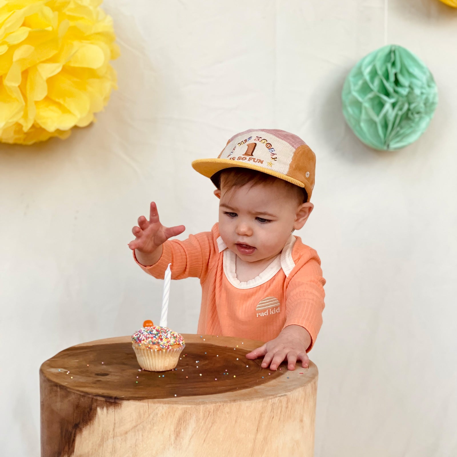 Party time photo, one year old child wearing Banabae Peach Ribbed Organic Cotton Long Sleeve Onesie with contrast natural neck bind and Rad kid/Banabae Sun Logo embroidered on right hand . Child is also wearing the 1st Birthday Cord Cap musk Spliced, she is reaching out to touch a birthday cupcake. party decorations in the background