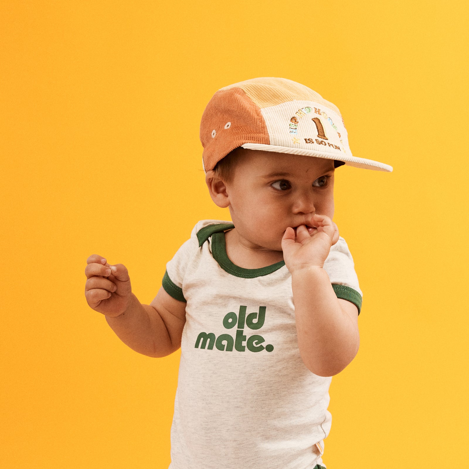 Child wearing the 1st Birthday Cord 5 Paneled Cap. which is a colour blocked design in blue, orange, natural and yellow . Front of cap is natural with HIP HIP HOORAY 1 IS SO FUN embroidered in multicolored thread. Blue peak with contrast Natural bind. They are also wearing an Old Mate Onesie which is light grey marl with Old Mate printed on the center front in green with contract green binding on neck and sleeves