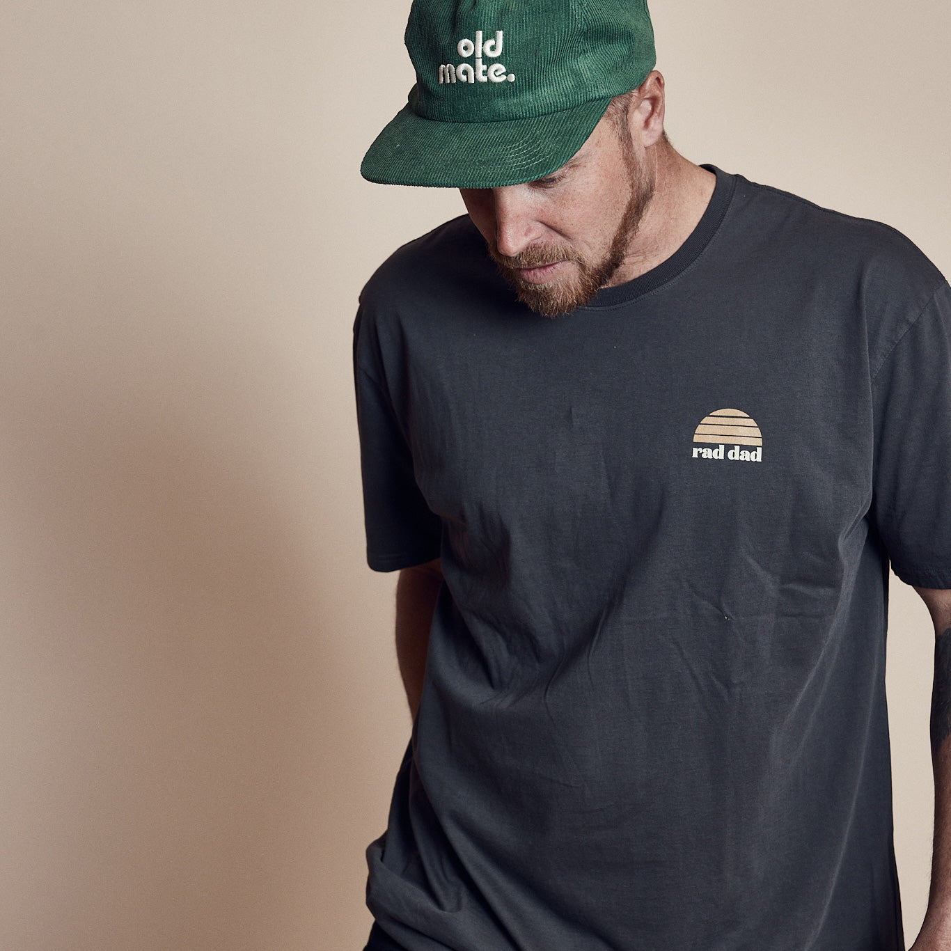 Man wearing a faded black organic cotton tee with a small "rad dad" logo on the upper left chest area and an "Old Mate' cap in green