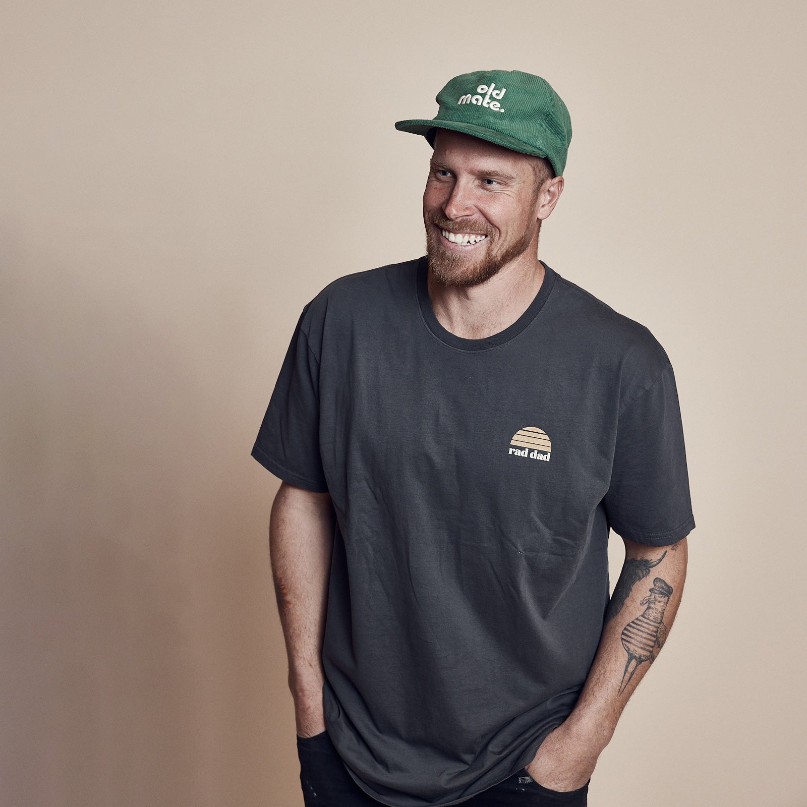 Man wearing a faded black organic cotton tee with a small "rad dad" logo on the upper left chest area and an "Old Mate' cap in green