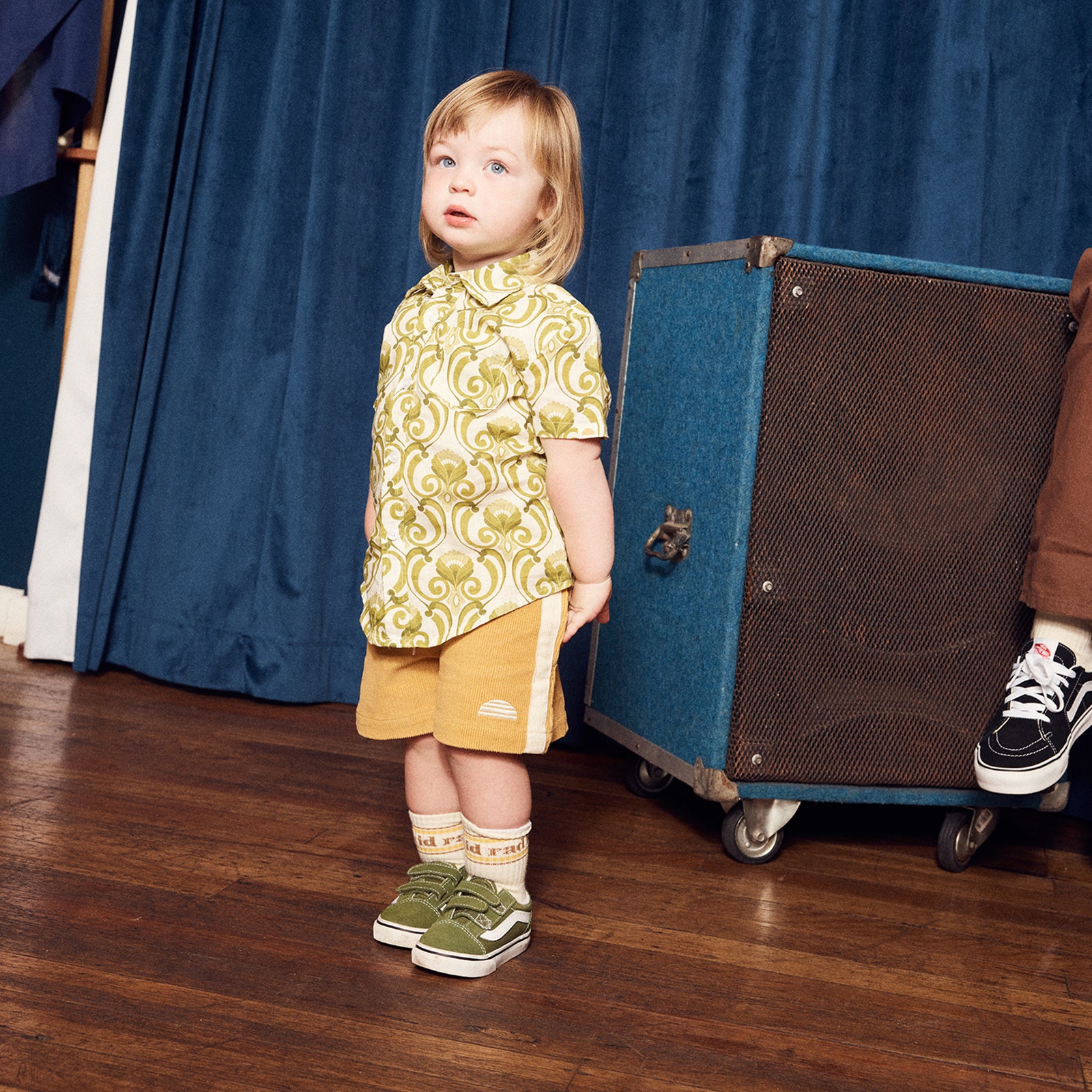 Child in a patterned shirt and yellow shorts standing next to a blue suitcase on a wooden floor.