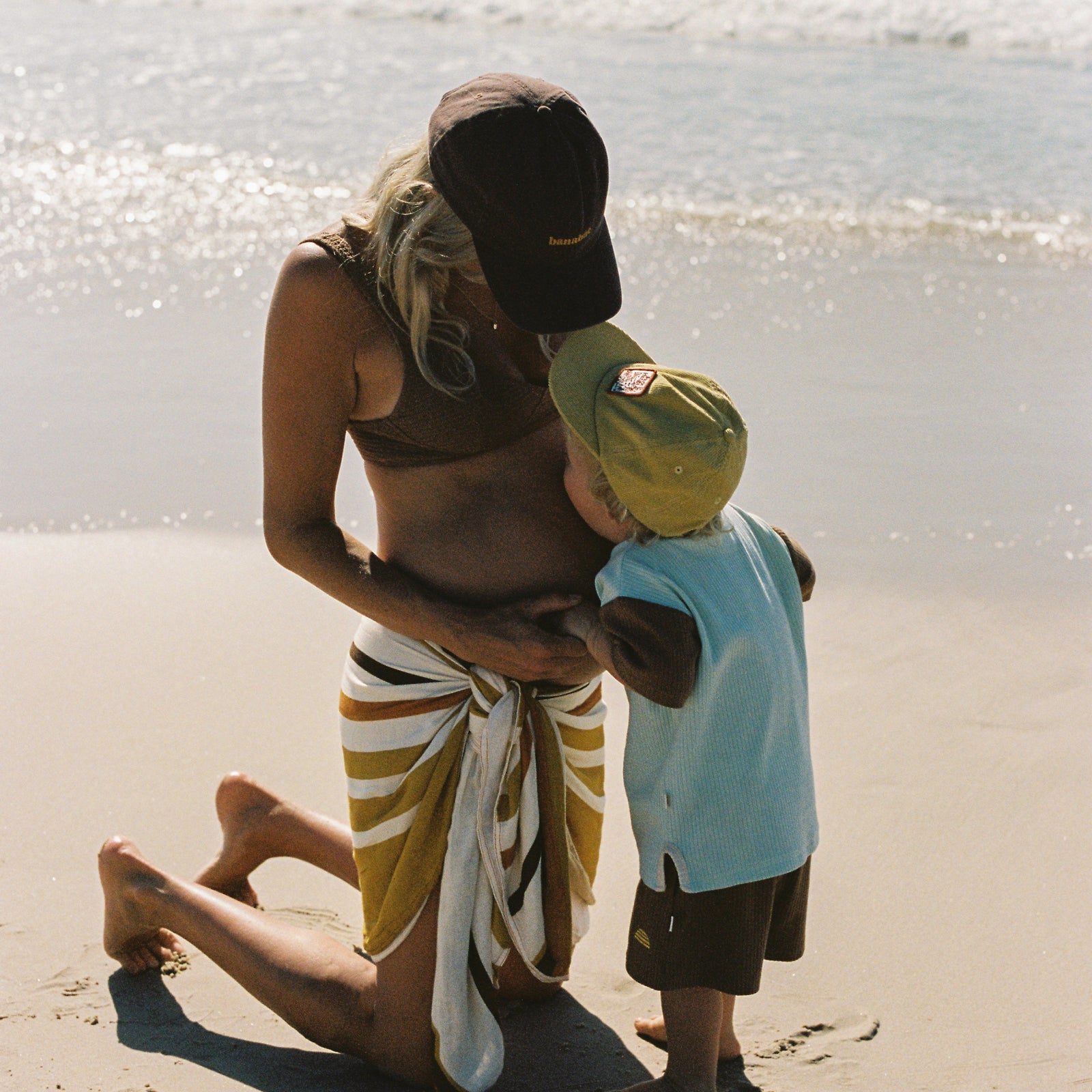 lifestyle image of pregnant women on the beach wearing a  brown bikini top, Banabae cap in brown with Banabae embroidered on the center front in yellow and Banabae Golden Daze Sarong tied around waist. Looking down at a small child wearing Banabae Park Hangs cap in yellow and Banabae ribbed shorts and Tee twin set in blue and brown, who is kissing her belly. 