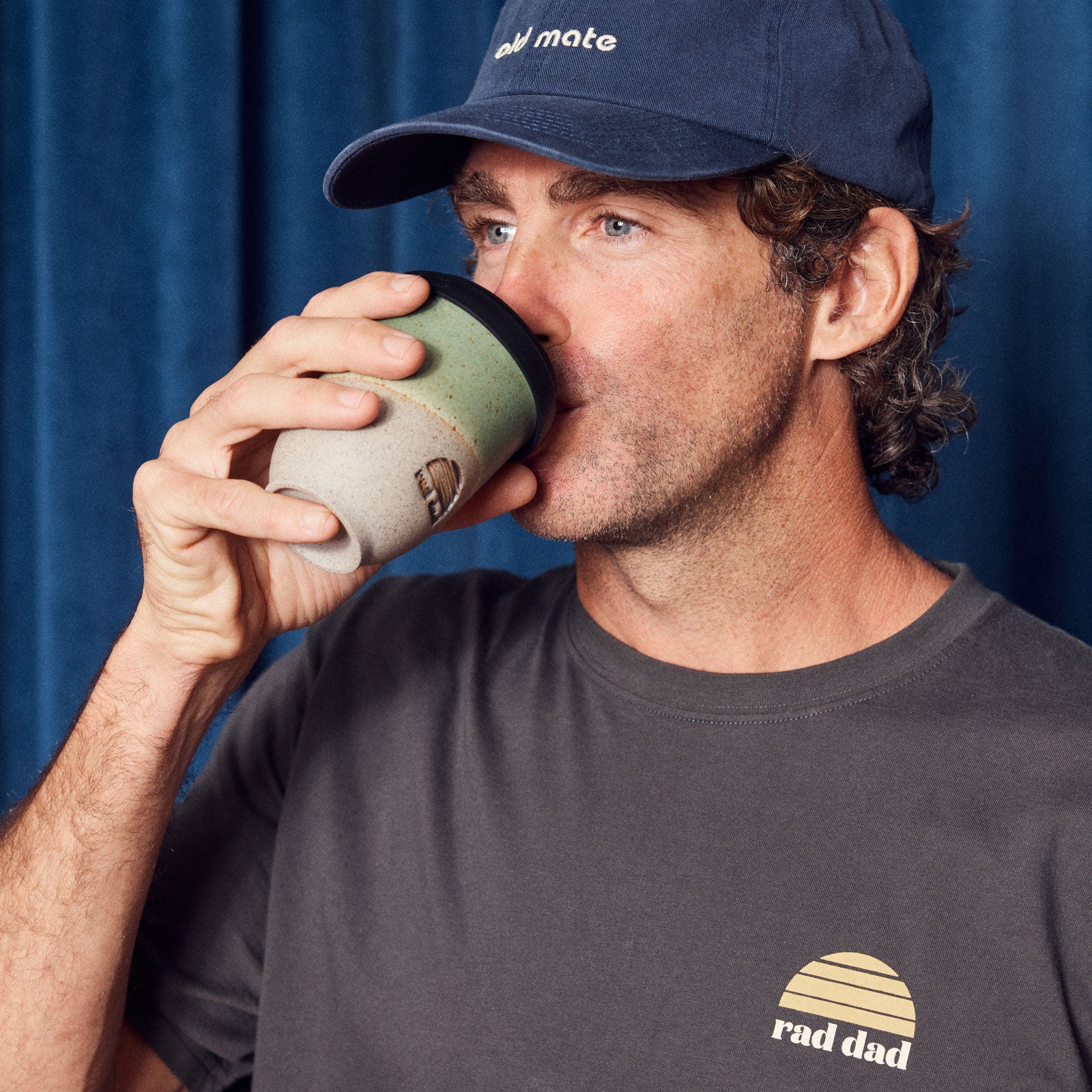 Man drinking from a coffee cup with a blue cap and 'rad dad' t-shirt against a blue background
