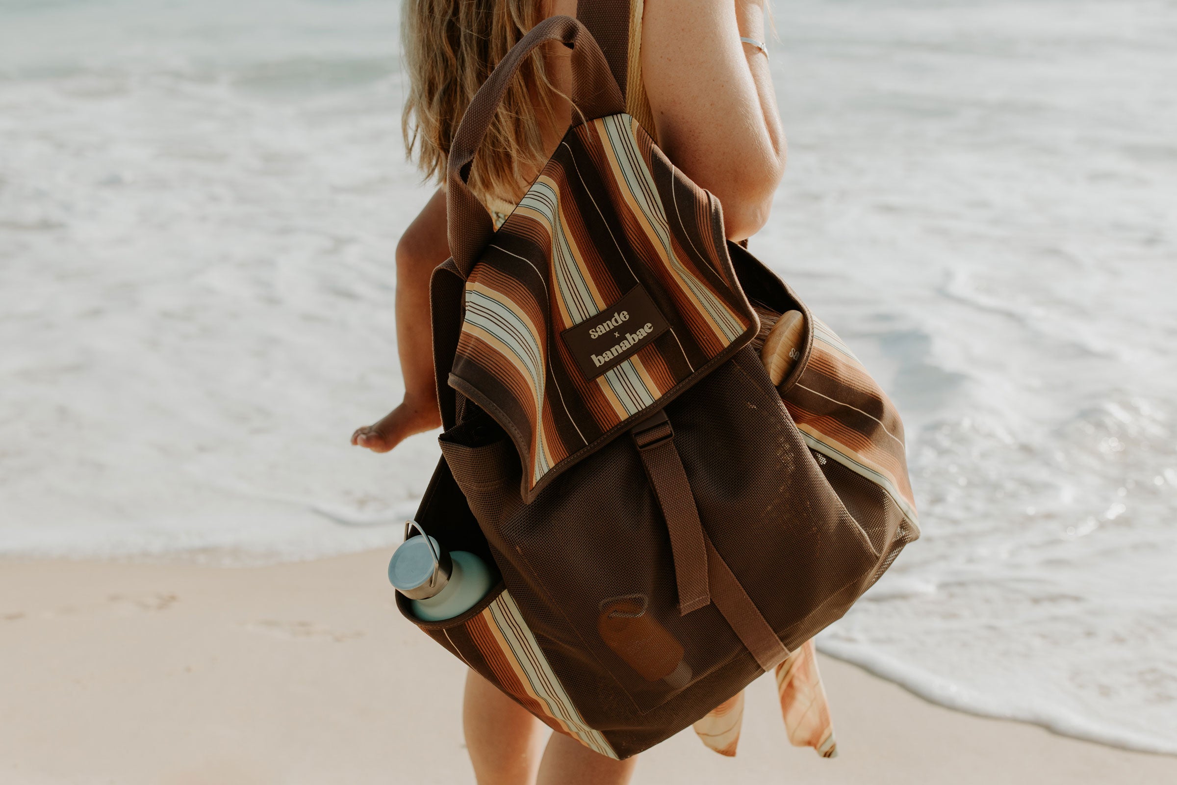 A woman on a beach carries a large brown and tan striped backpack with a small bottle in the side pocket. The backpack has a visible patch reading 'Sande x Banabae'.